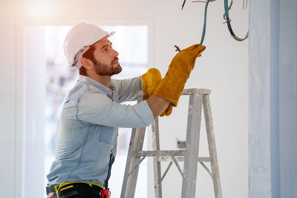 Bearded man in hard hat and protective gloves on a ladder inspecting ceiling wiring