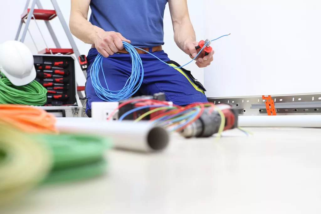 Man snipping end of coiled wire while kneeling among electrical tools on floor.