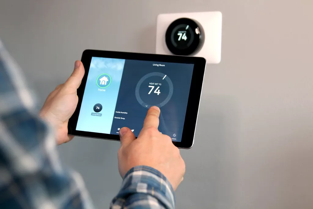 Man's hands holding a tablet and adjusting temp on his smart thermostat, with the thermostat in the background. 