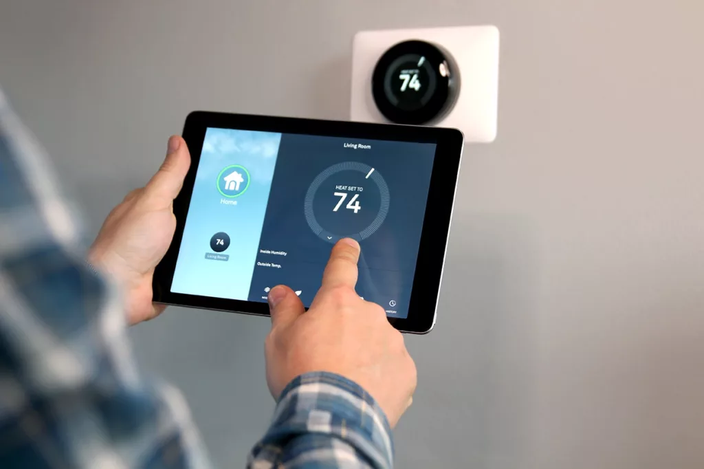 Man's hands holding a tablet and adjusting temp on his smart thermostat, with the thermostat in the background.