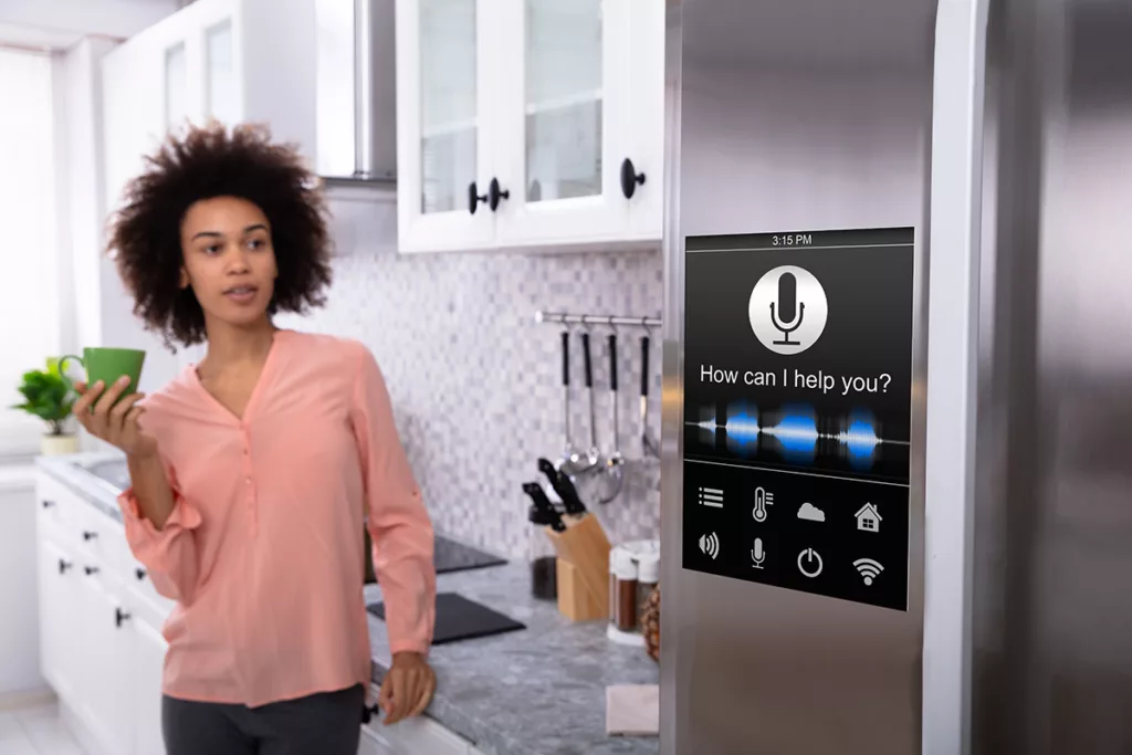 Woman holding coffee cup in kitchen with voice-command smart refrigerator asking "How can I help you?"