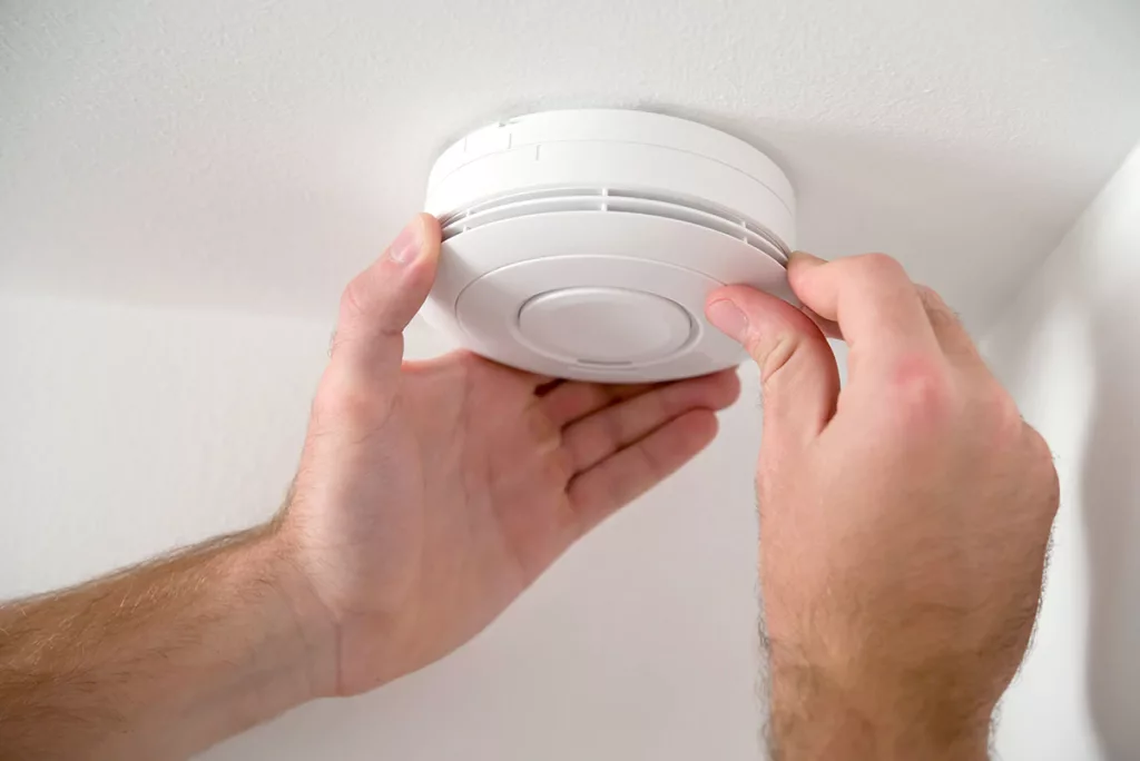 An electrician installs a smoke detector on a white ceiling for safety.