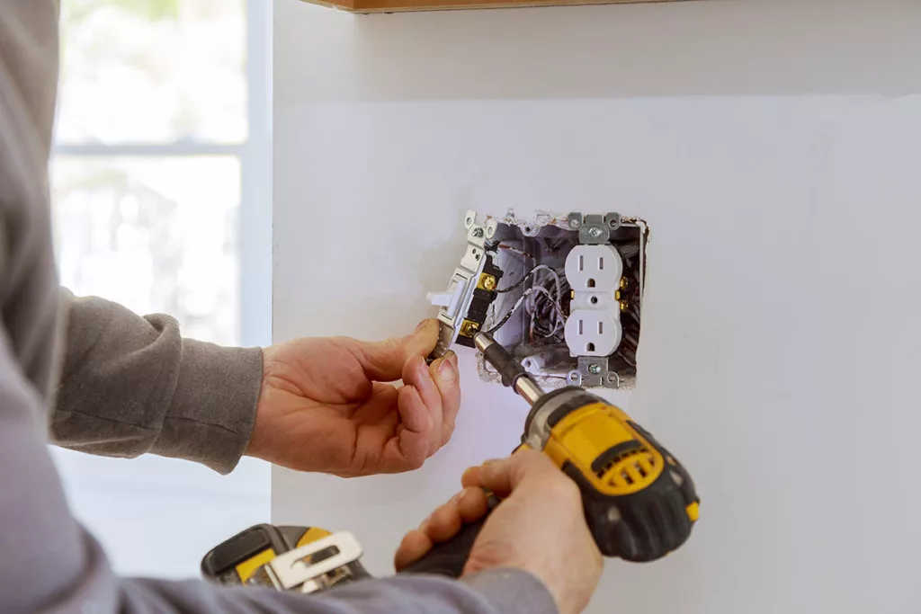 An electrician repairs an electrical outlet for home safety, with a white wall in the background.