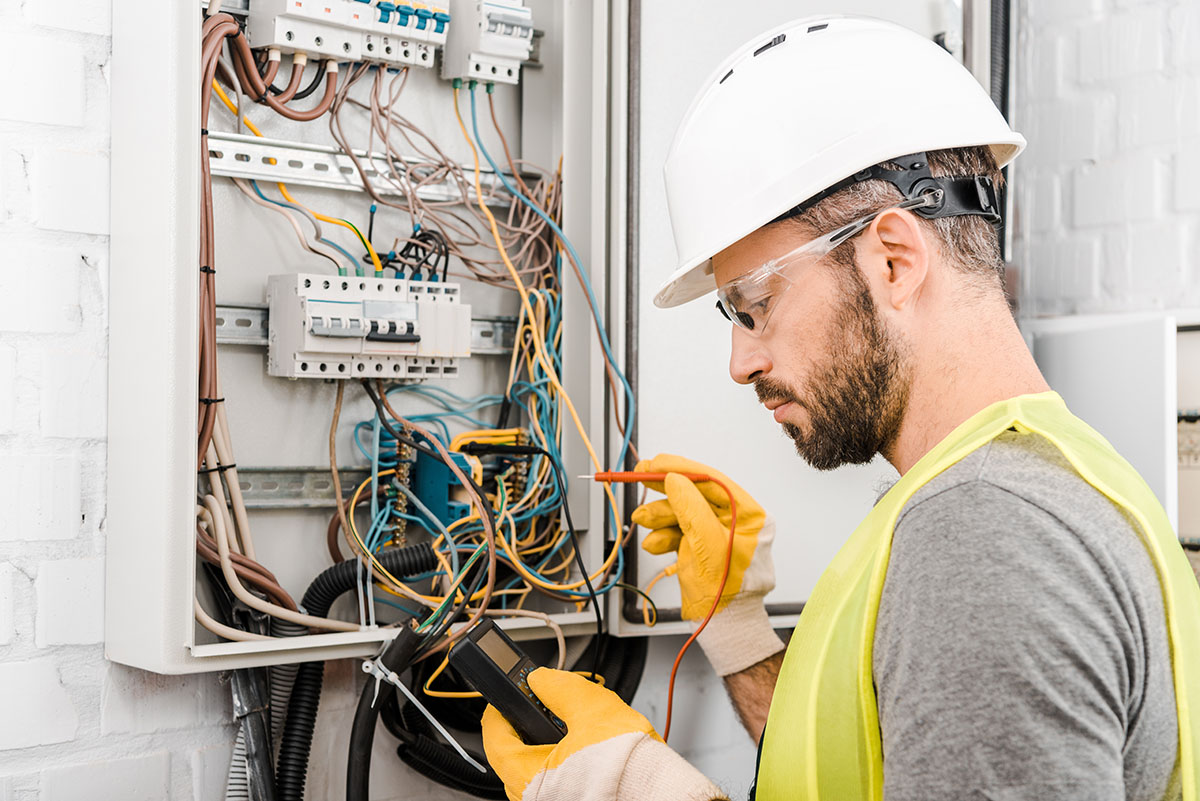 A male electrician with a hard hat inspects an electrical panel for safety.