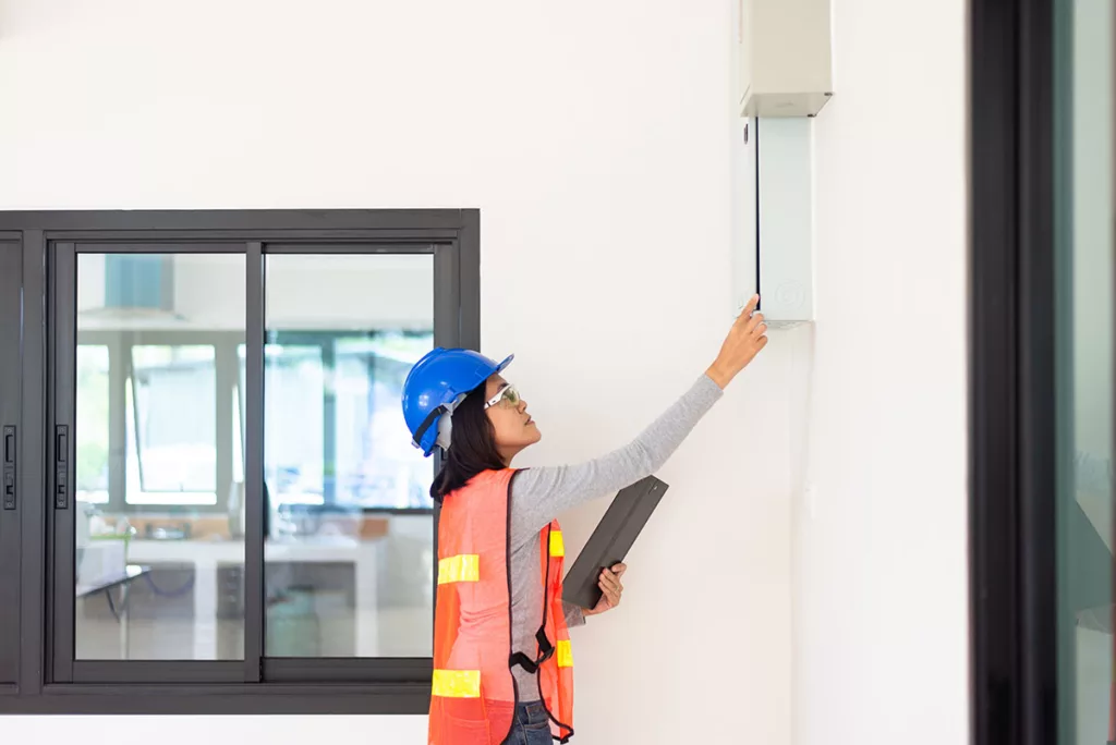 A woman wearing a hard hat and orange vest inspects the electrical safety of a home.