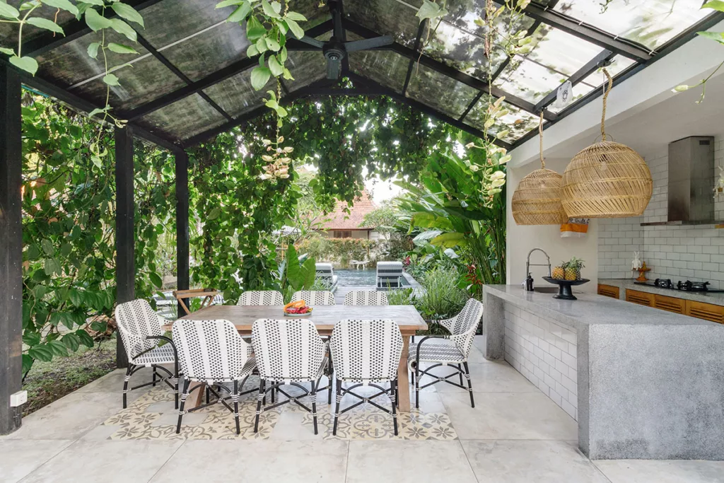 An outdoor kitchen with wicker pendant lights, and a dining table with chairs against a background of plants