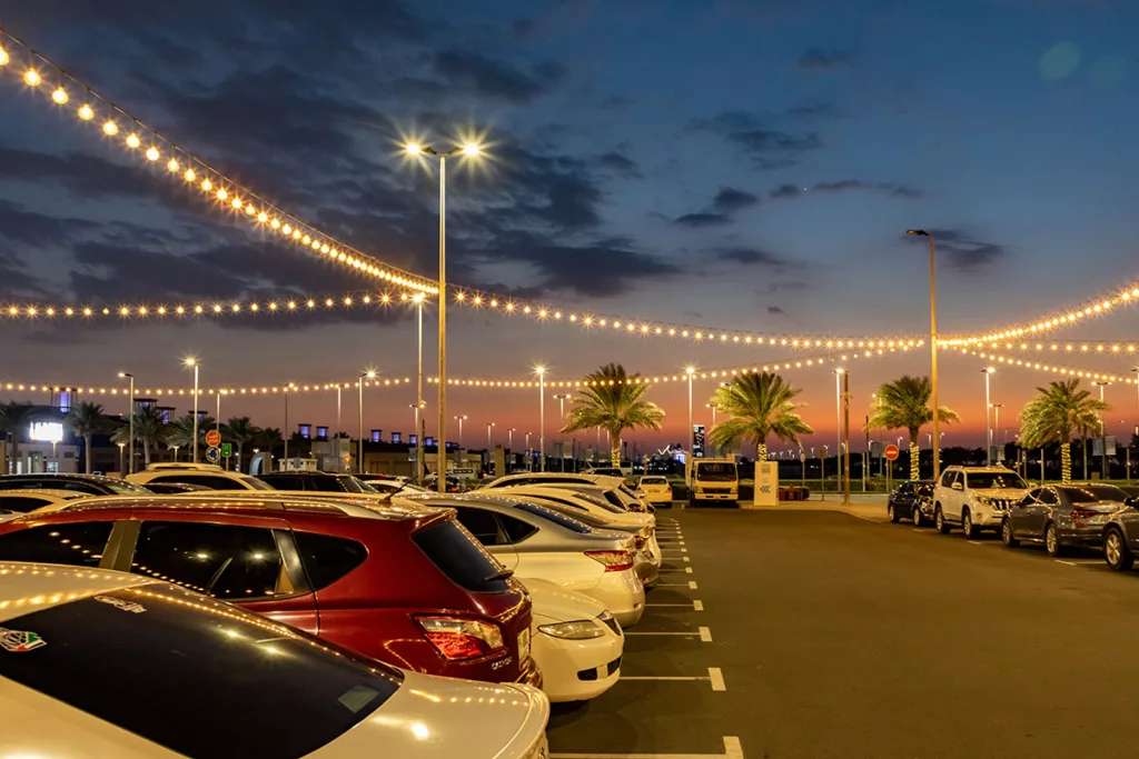 A commercial parking lot at sunset, filled with cars, street lamps, palm trees, and string lights.