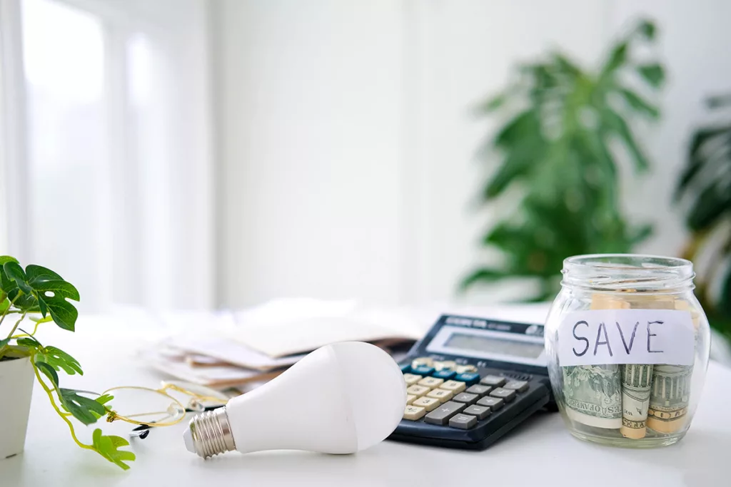 An LED light bulb on a white table, next to a calculator and a savings jar of money