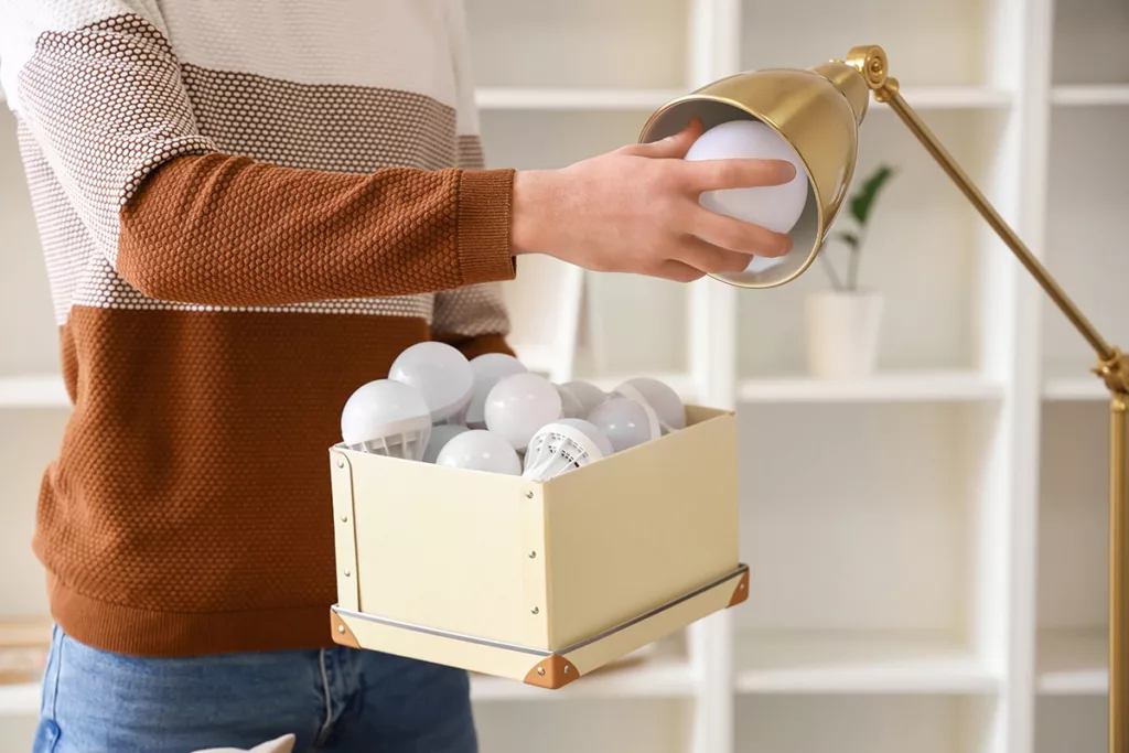 A homeowner holds a box of LED lightbulbs and changes the lightbulb of a gold lamp.