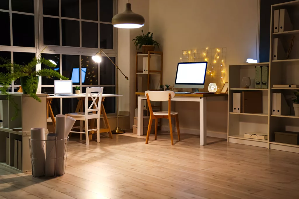 A home office at night with wood floors, plants, a desk with a computer, and bright light fixtures with warm LED light bulbs