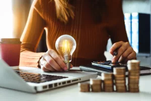 A woman sits at a table in front of a laptop, holding an LED light bulb and calculating savings from a lowered electricity bill.