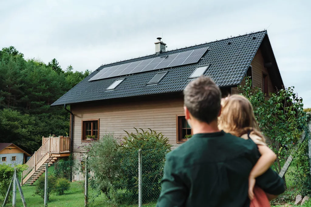 Rear view of dad holding her little girl in arms and looking at their house with solar panels.