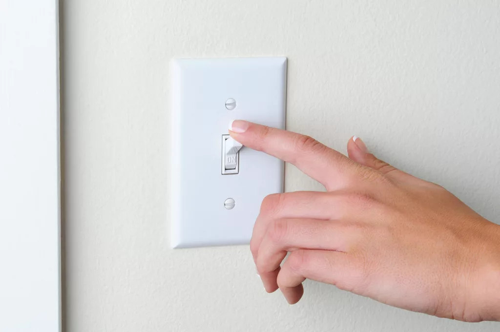 A close up of a hand turning off a light switch to save energy and lower their electricity bill