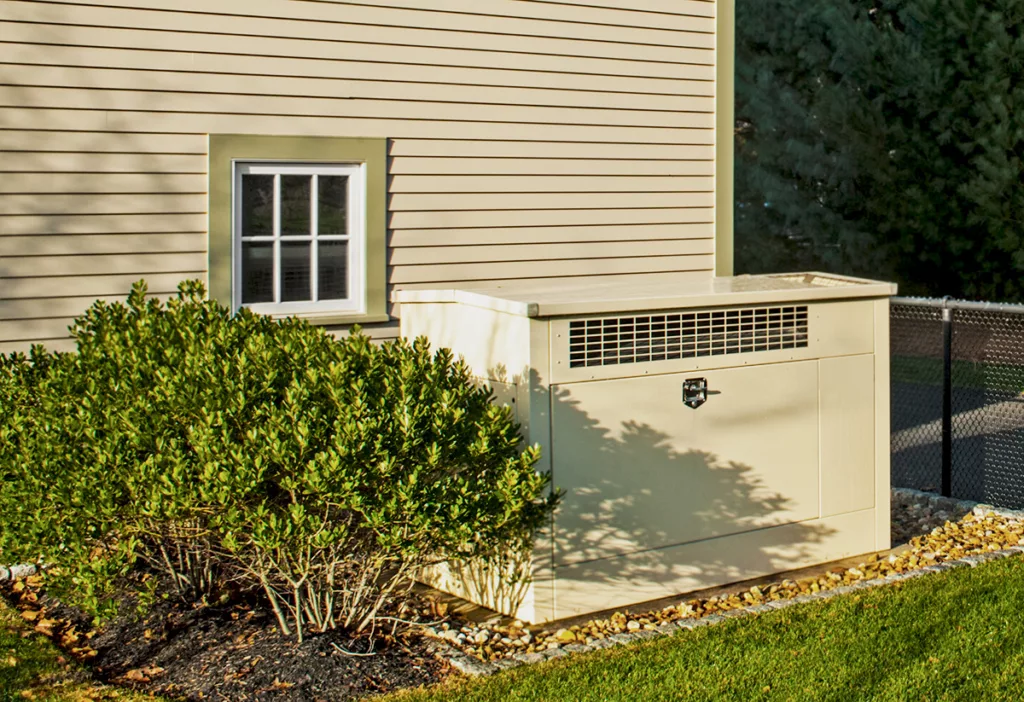 A residential standby generator in the backyard of a house with beige walls.
