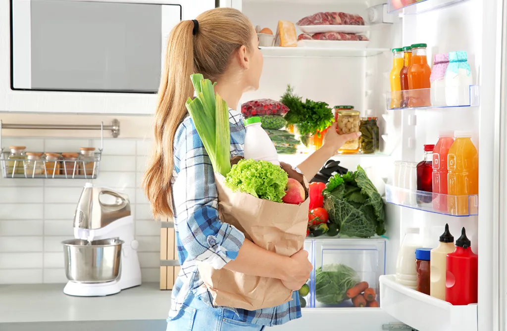 A woman puts away groceries in an open refrigerator in a bright kitchen.