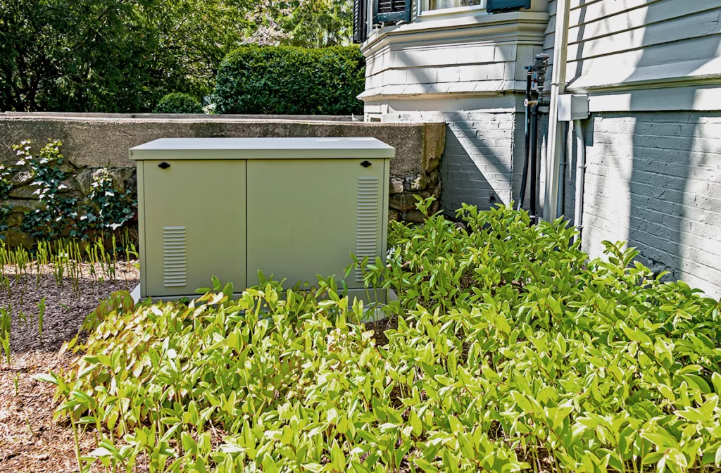 A residential standby generator in the backyard of a house, surrounded by green plants.