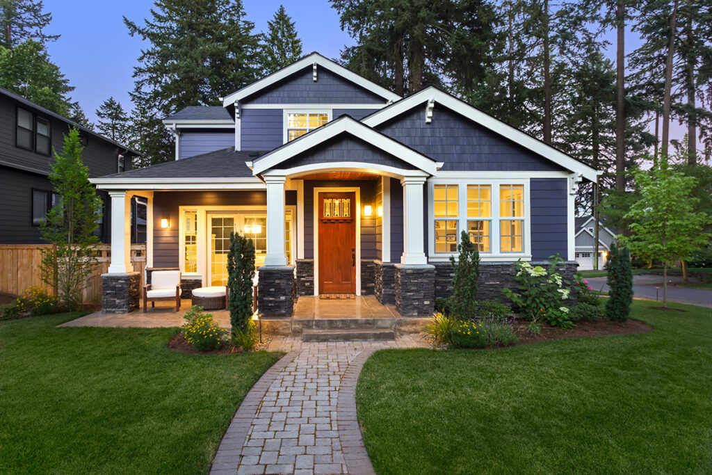 The exterior of a dark blue house in the evening, with glowing front porch lights, a big green lawn, and trees.