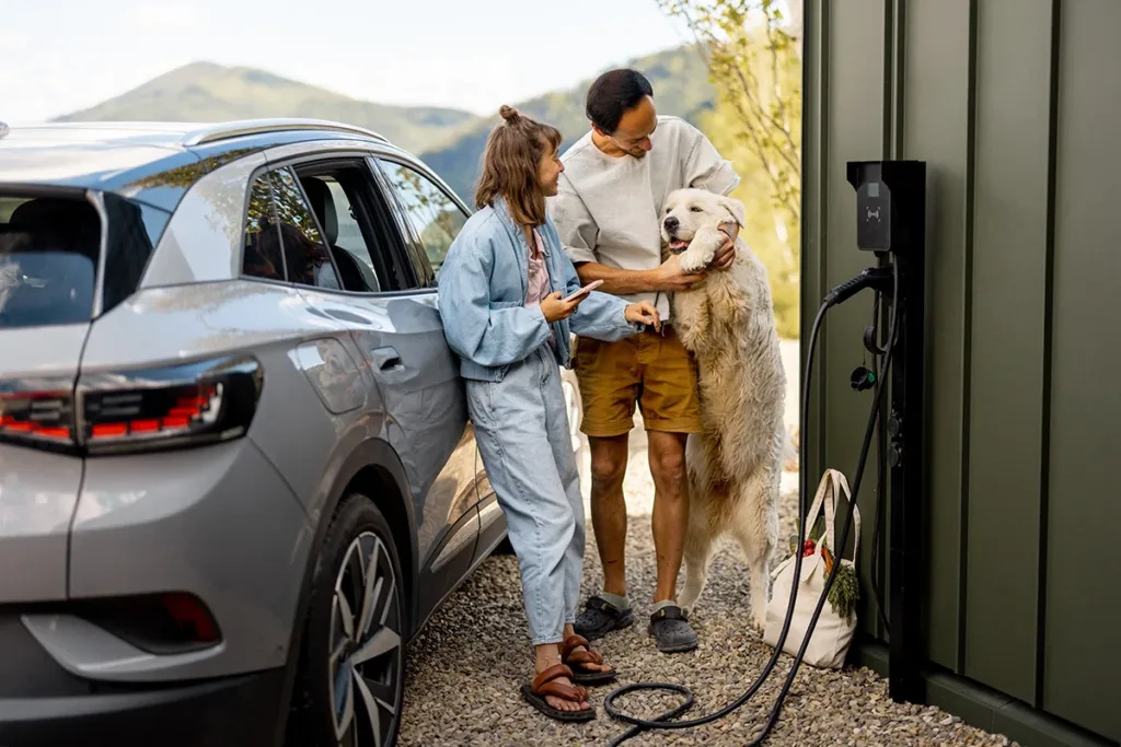 A young couple with a dog stands by their home's EV charger, waiting for their gray electric vehicle to charge