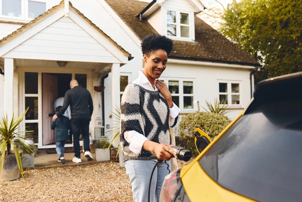 A homeowner standing in front of her house, next to a yellow electric vehicle, plugging in an EV charger